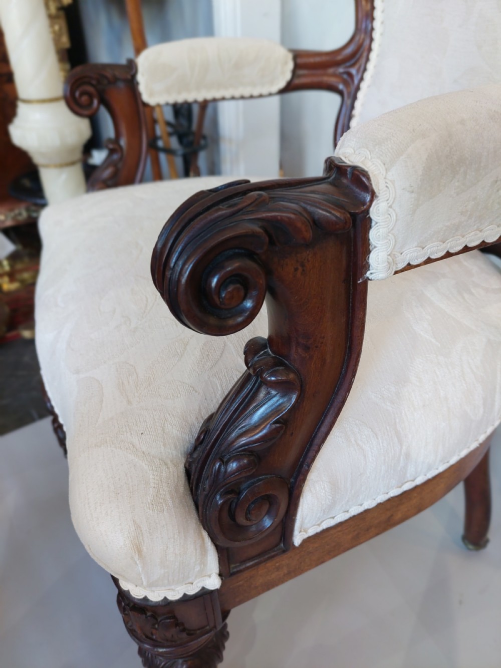 An antique Regency carved mahogany armchair upholstered in cream damask fabric, featuring scroll arms and turned front legs with castors. View II