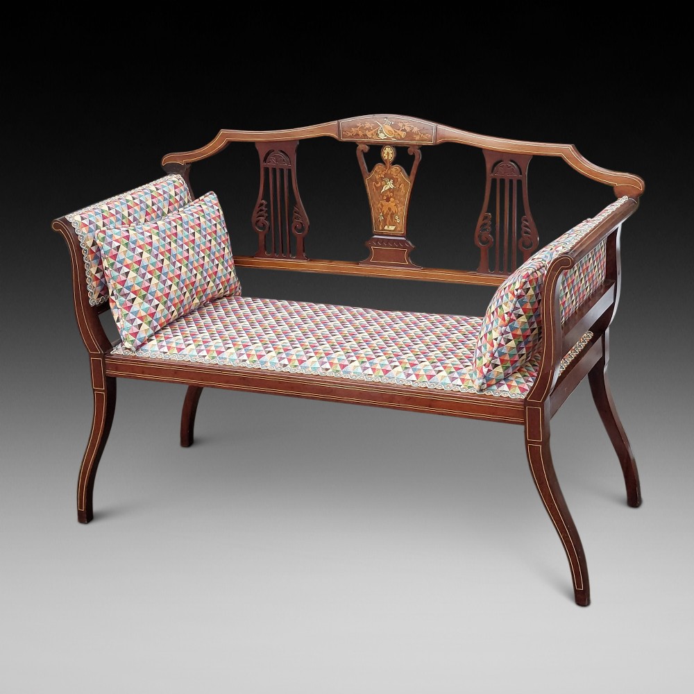 An Edwardian mahogany two-seater settee with decorative central marquetry splats, curved arms, splayed legs, and colourful geometric upholstery.