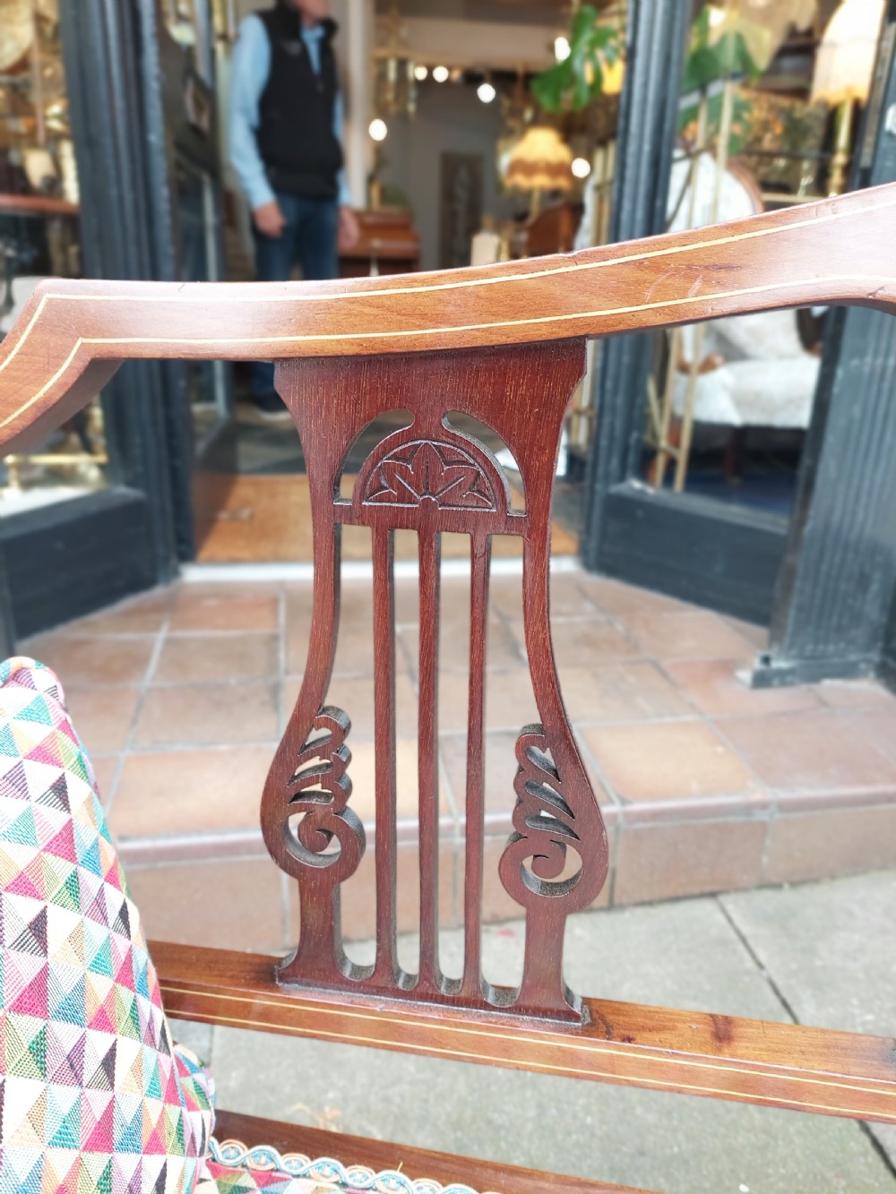 An Edwardian mahogany two-seater settee with decorative central marquetry splats, curved arms, splayed legs, and colourful geometric upholstery. View III