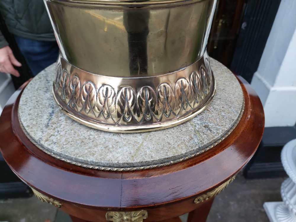 A polished Edwardian brass half-moon stick stand featuring embossed decorative borders and a dark wooden carrying handle. View III