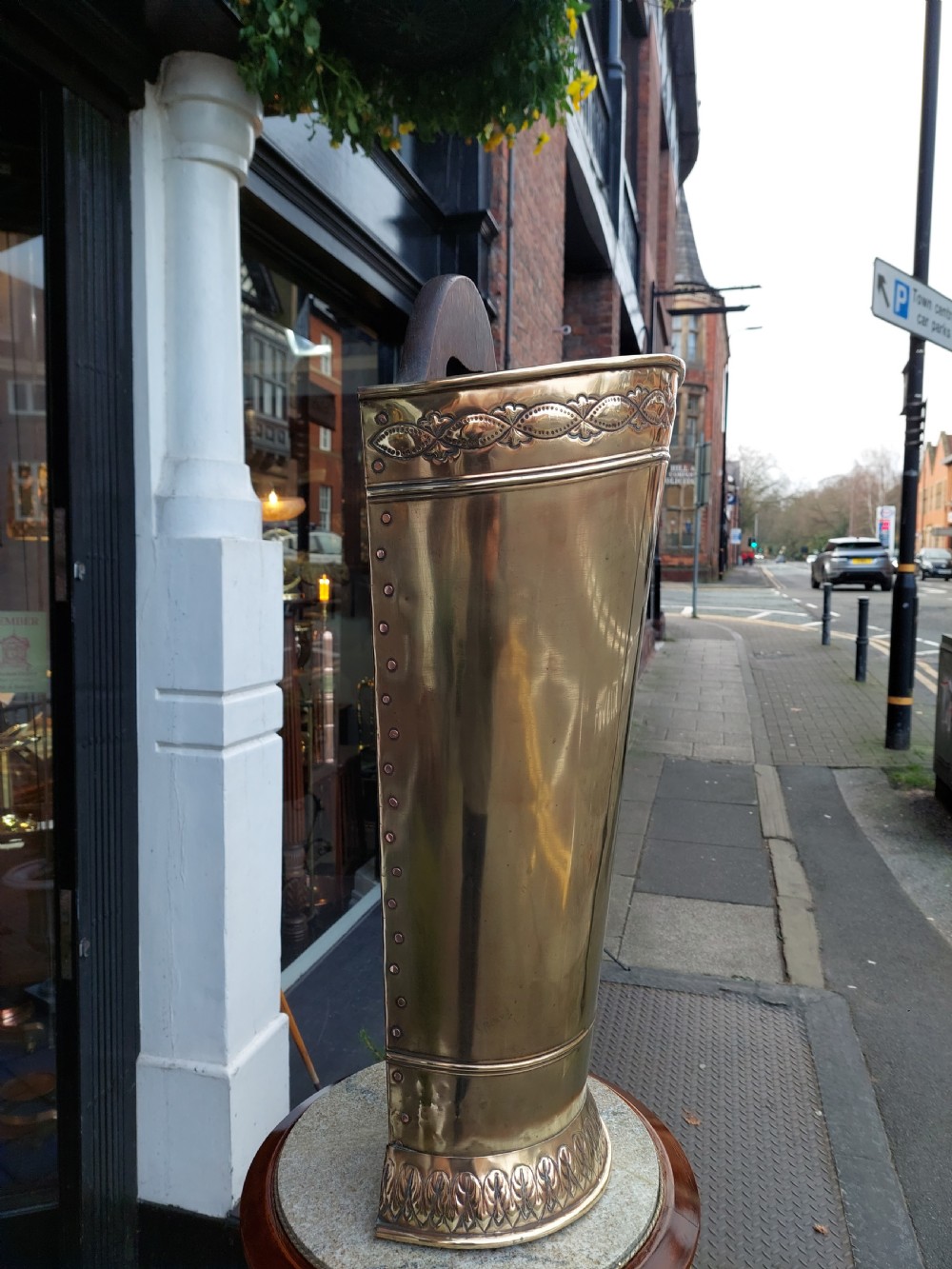 A polished Edwardian brass half-moon stick stand featuring embossed decorative borders and a dark wooden carrying handle. View VI