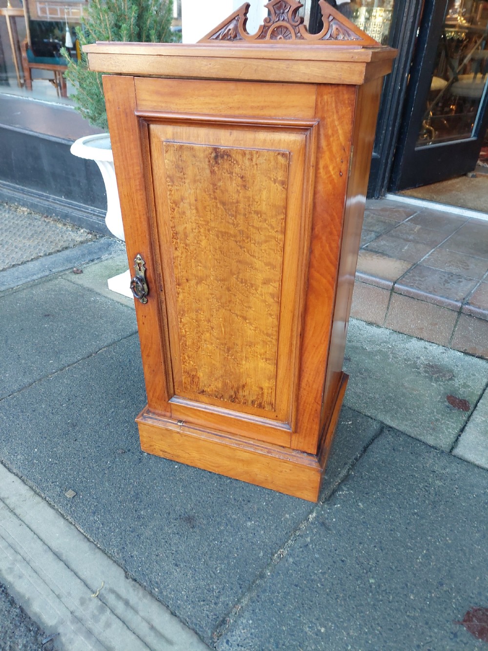 An Edwardian walnut bedside cabinet featuring a single cupboard door with a figured wood panel, a carved top gallery, and a solid plinth base. View IV