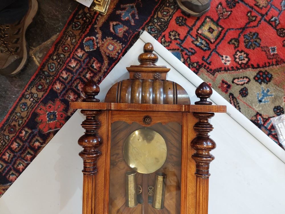 Late Victorian double weight Vienna wall clock featuring a carved walnut case topped with an eagle pediment, turned side finials, a glazed door revealing twin brass weights, and a white dial with Roman numerals. View II