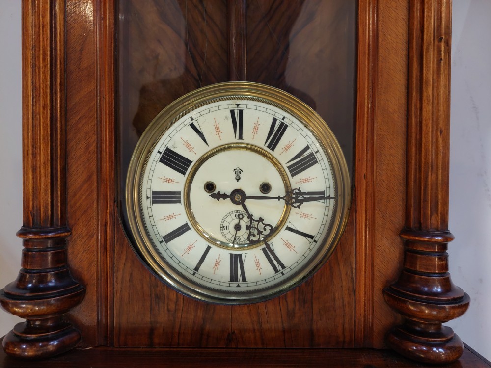 Late Victorian double weight Vienna wall clock featuring a carved walnut case topped with an eagle pediment, turned side finials, a glazed door revealing twin brass weights, and a white dial with Roman numerals. View III