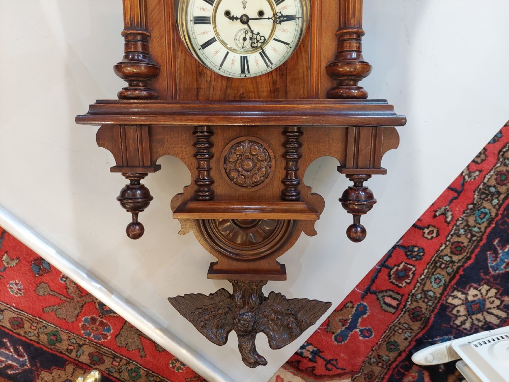 Late Victorian double weight Vienna wall clock featuring a carved walnut case topped with an eagle pediment, turned side finials, a glazed door revealing twin brass weights, and a white dial with Roman numerals. View IV