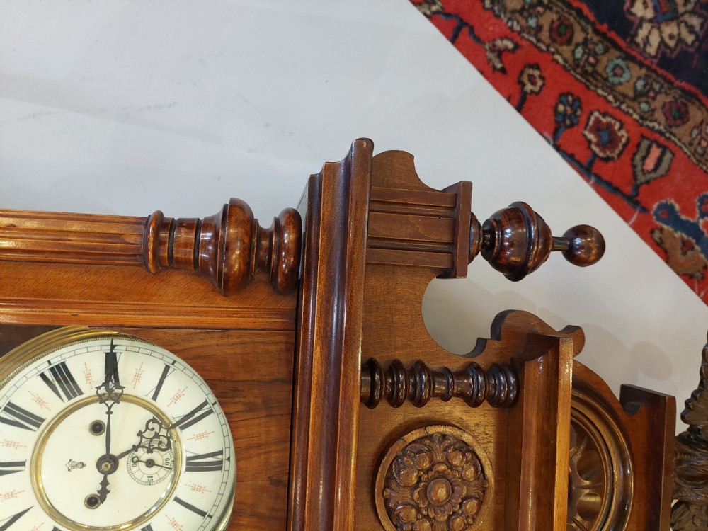 Late Victorian double weight Vienna wall clock featuring a carved walnut case topped with an eagle pediment, turned side finials, a glazed door revealing twin brass weights, and a white dial with Roman numerals. View V
