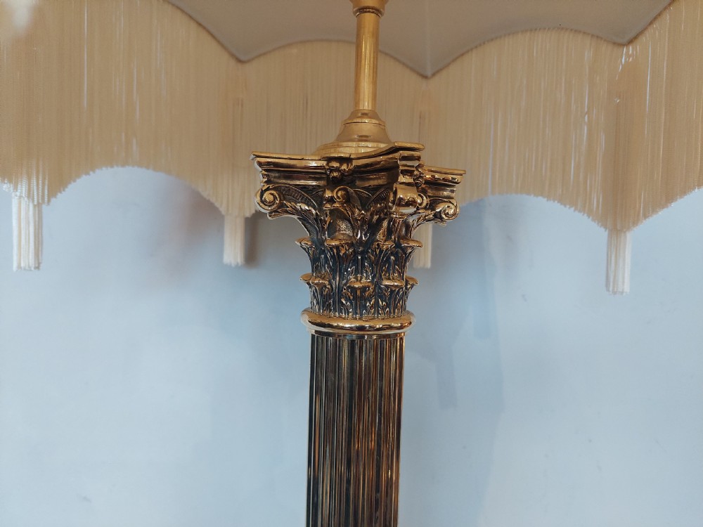 An Edwardian brass table lamp with a fluted Corinthian column base, topped with a cream patterned fabric shade with long fringe trim. View IV
