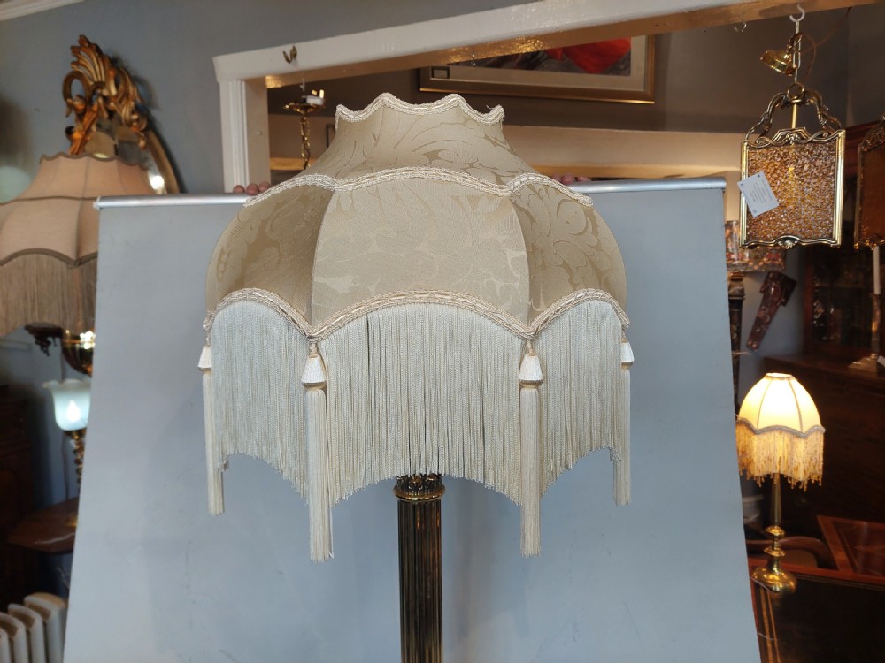 An Edwardian brass table lamp with a fluted Corinthian column base, topped with a cream patterned fabric shade with long fringe trim. View V