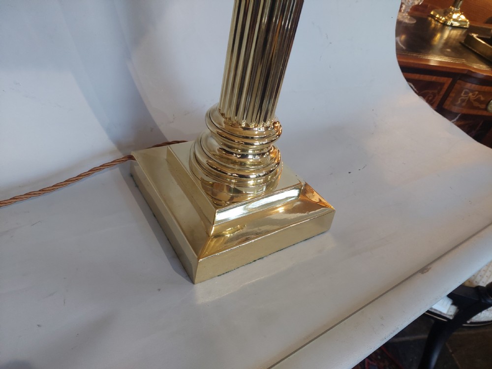 An Edwardian brass table lamp with a fluted Corinthian column base, topped with a cream patterned fabric shade with long fringe trim. View VI
