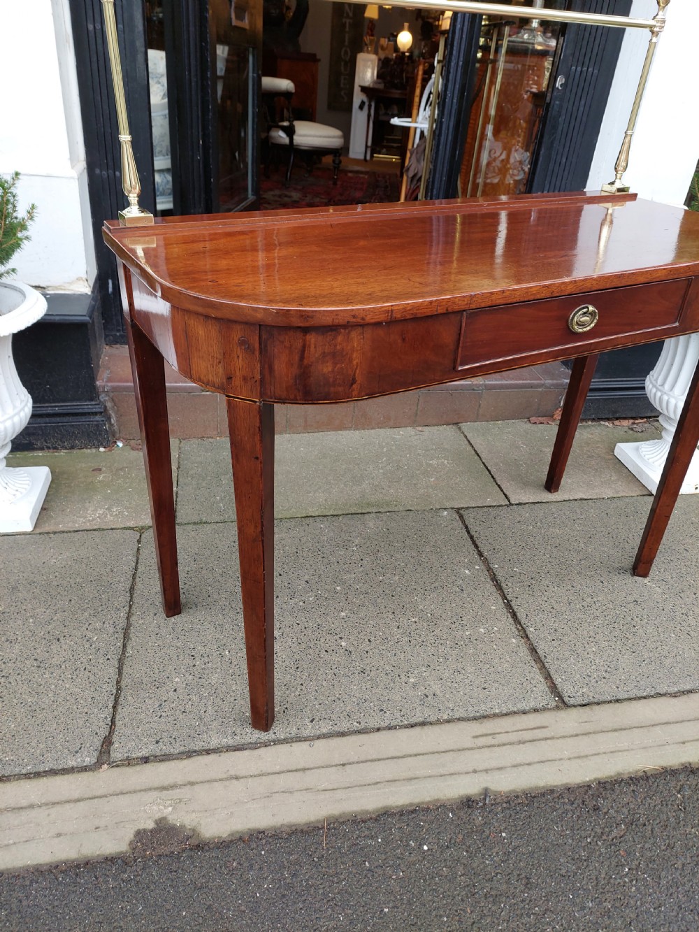 A Georgian mahogany hall table with a rounded bowfront top, a single central drawer with brass handle, a brass finial gallery rail, and four square tapered legs. View V