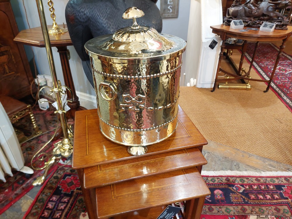 A cylindrical Victorian brass coal receiver featuring embossed garland motifs, beaded horizontal bands, side ring handles, and a fitted domed lid with a finial. View II
