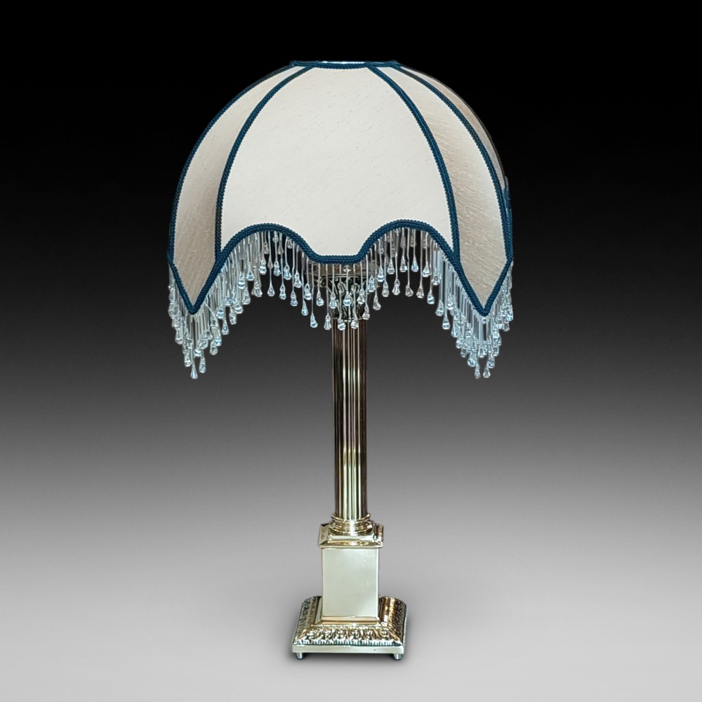 A brass column table lamp featuring a reeded stem, stepped square base, and a cream fabric dome shade trimmed with dark braid and clear glass droplet fringe.