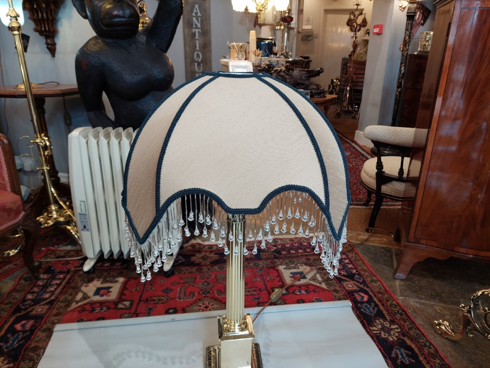 A brass column table lamp featuring a reeded stem, stepped square base, and a cream fabric dome shade trimmed with dark braid and clear glass droplet fringe. View VI
