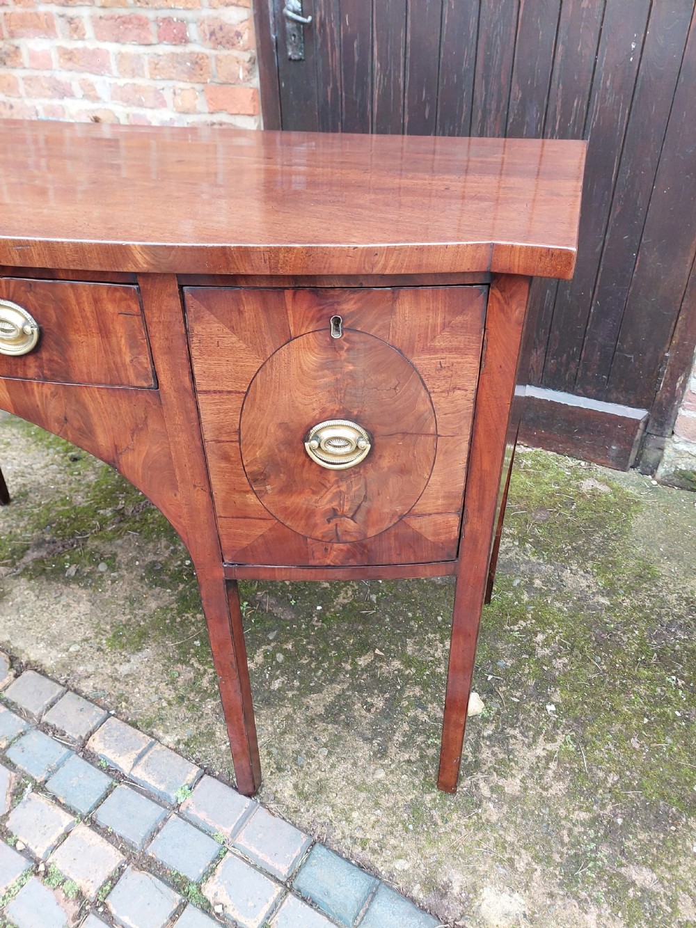 A late 18th-century mahogany bowfront sideboard featuring an arched central apron, oval-patterned veneered drawer fronts, brass drop handles, and six square tapered legs. View III