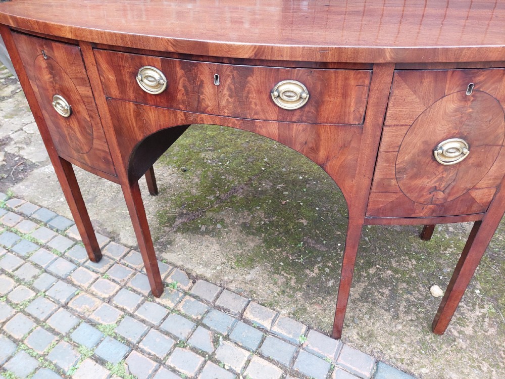 A late 18th-century mahogany bowfront sideboard featuring an arched central apron, oval-patterned veneered drawer fronts, brass drop handles, and six square tapered legs. View V