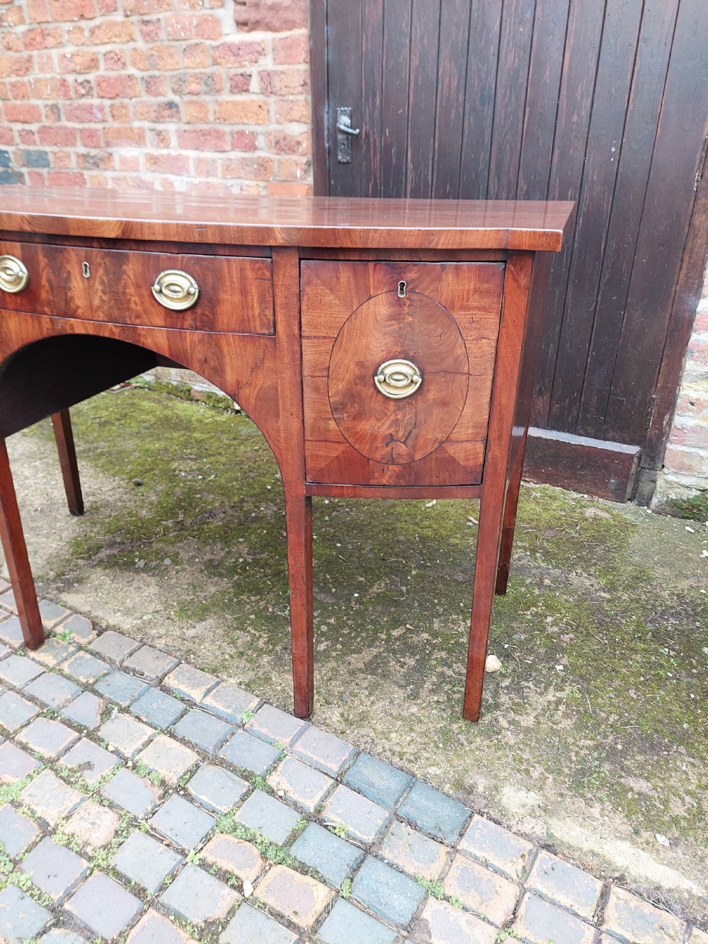 A late 18th-century mahogany bowfront sideboard featuring an arched central apron, oval-patterned veneered drawer fronts, brass drop handles, and six square tapered legs. View VI