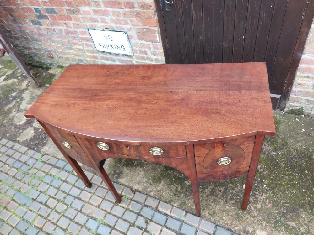 A late 18th-century mahogany bowfront sideboard featuring an arched central apron, oval-patterned veneered drawer fronts, brass drop handles, and six square tapered legs. View VII