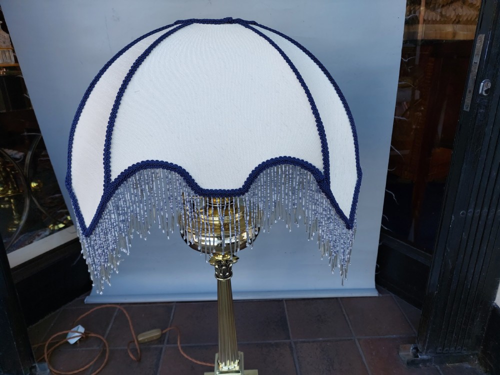 A late nineteenth-century Corinthian column oil lamp featuring a fluted stem, polished metal reservoir, and a white fabric dome shade trimmed with blue piping and long glass bead fringe. View II
