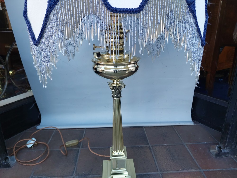A late nineteenth-century Corinthian column oil lamp featuring a fluted stem, polished metal reservoir, and a white fabric dome shade trimmed with blue piping and long glass bead fringe. View III