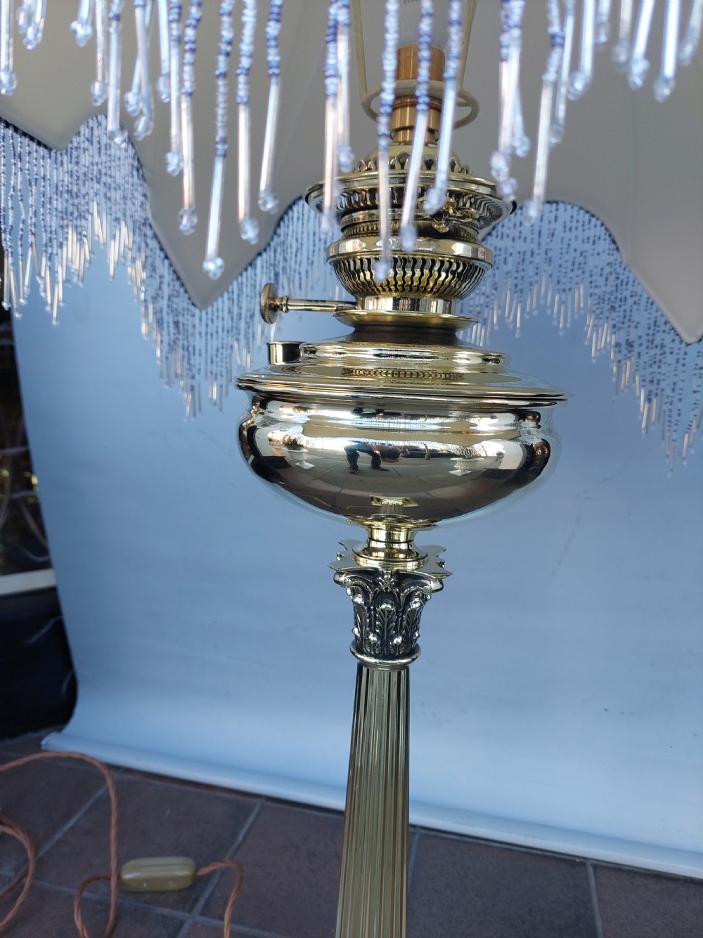 A late nineteenth-century Corinthian column oil lamp featuring a fluted stem, polished metal reservoir, and a white fabric dome shade trimmed with blue piping and long glass bead fringe. View IV