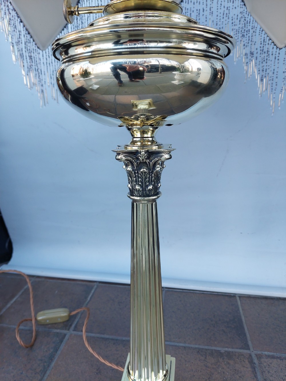 A late nineteenth-century Corinthian column oil lamp featuring a fluted stem, polished metal reservoir, and a white fabric dome shade trimmed with blue piping and long glass bead fringe. View V