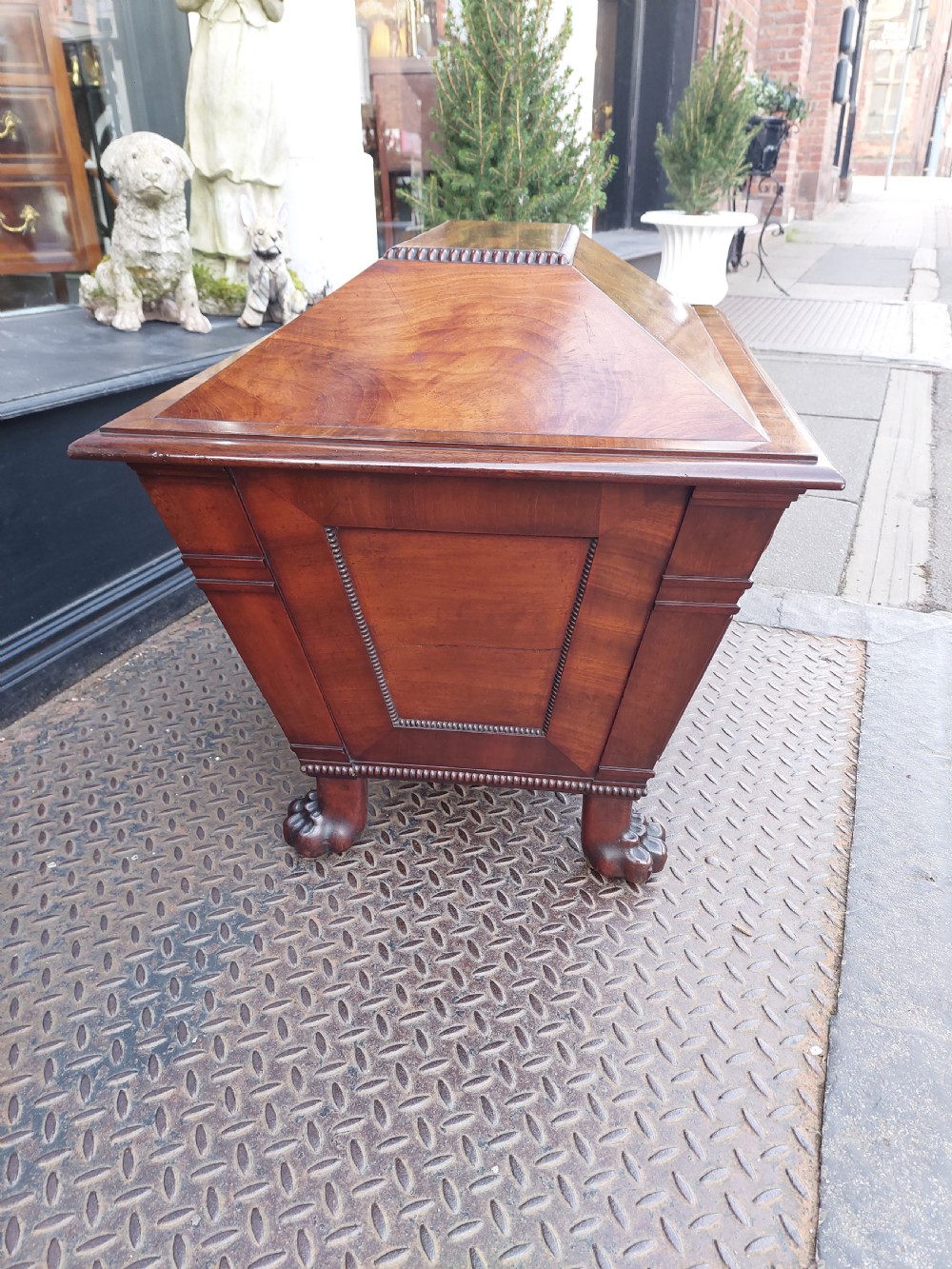 A late Regency flame mahogany sarcophagus cellarette featuring a faceted hinged lid, carved lion paw feet, and beaded edge moulding. View V