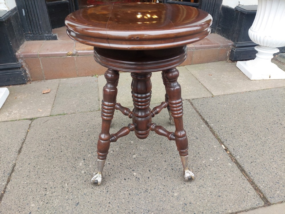 An early 20th-century dark mahogany piano stool featuring a circular swivel top, turned spindled legs, central pillar, and metal claw feet with glass balls. View II
