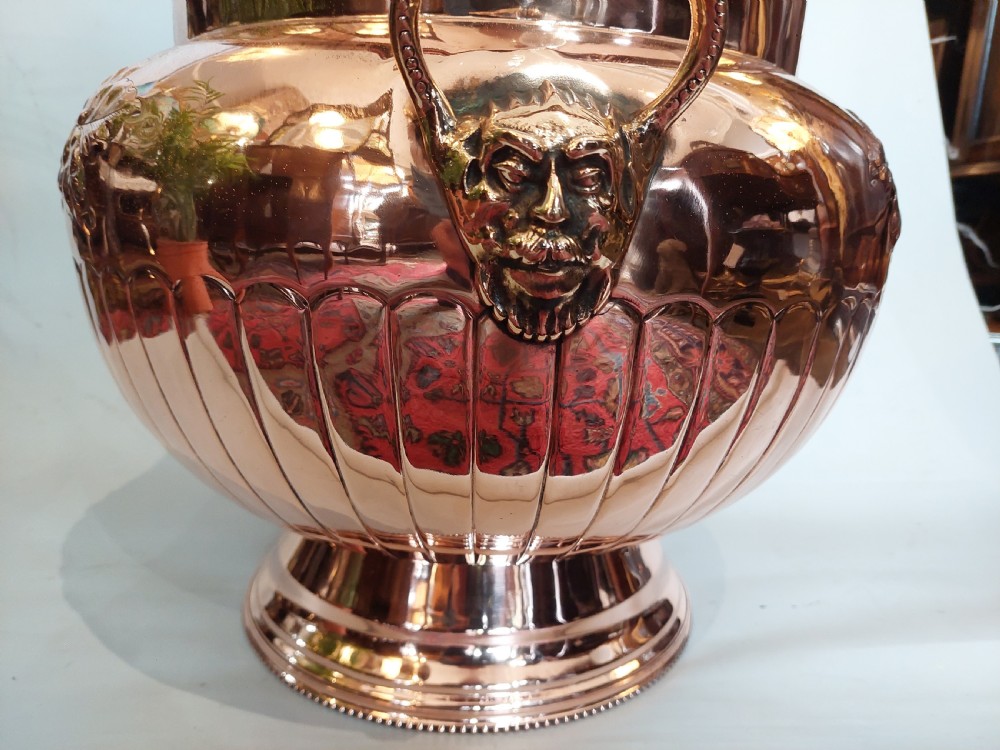 A bulbous Victorian polished copper vessel featuring embossed floral scrollwork, lower gadrooned fluting, two side loop handles, and a circular stepped pedestal base. View V