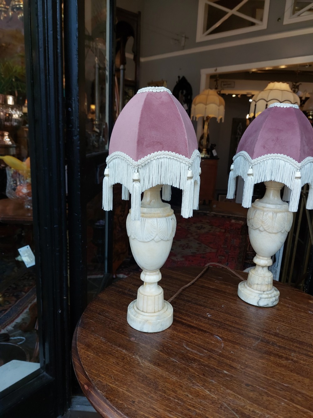 A pair of carved white alabaster urn-form table lamps featuring pink velvet shades trimmed with white tassel fringe. View II