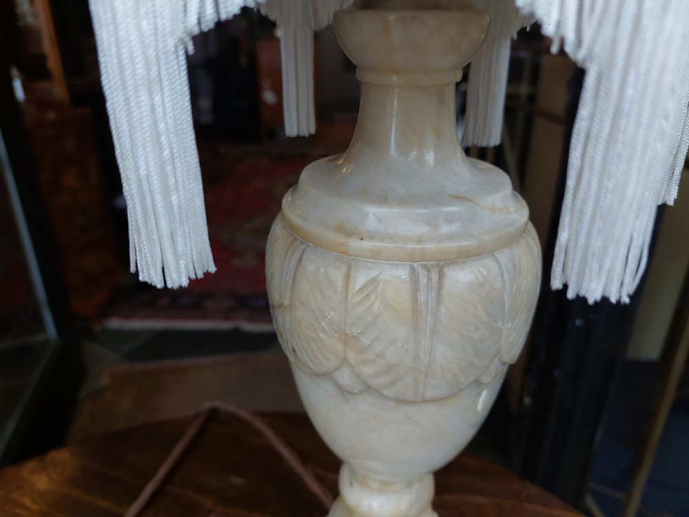 A pair of carved white alabaster urn-form table lamps featuring pink velvet shades trimmed with white tassel fringe. View IV