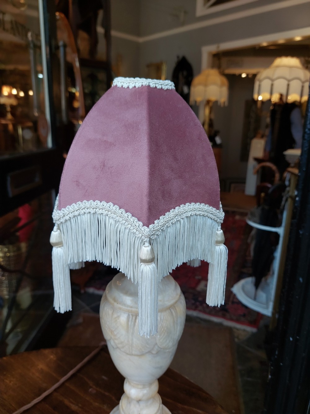 A pair of carved white alabaster urn-form table lamps featuring pink velvet shades trimmed with white tassel fringe. View V