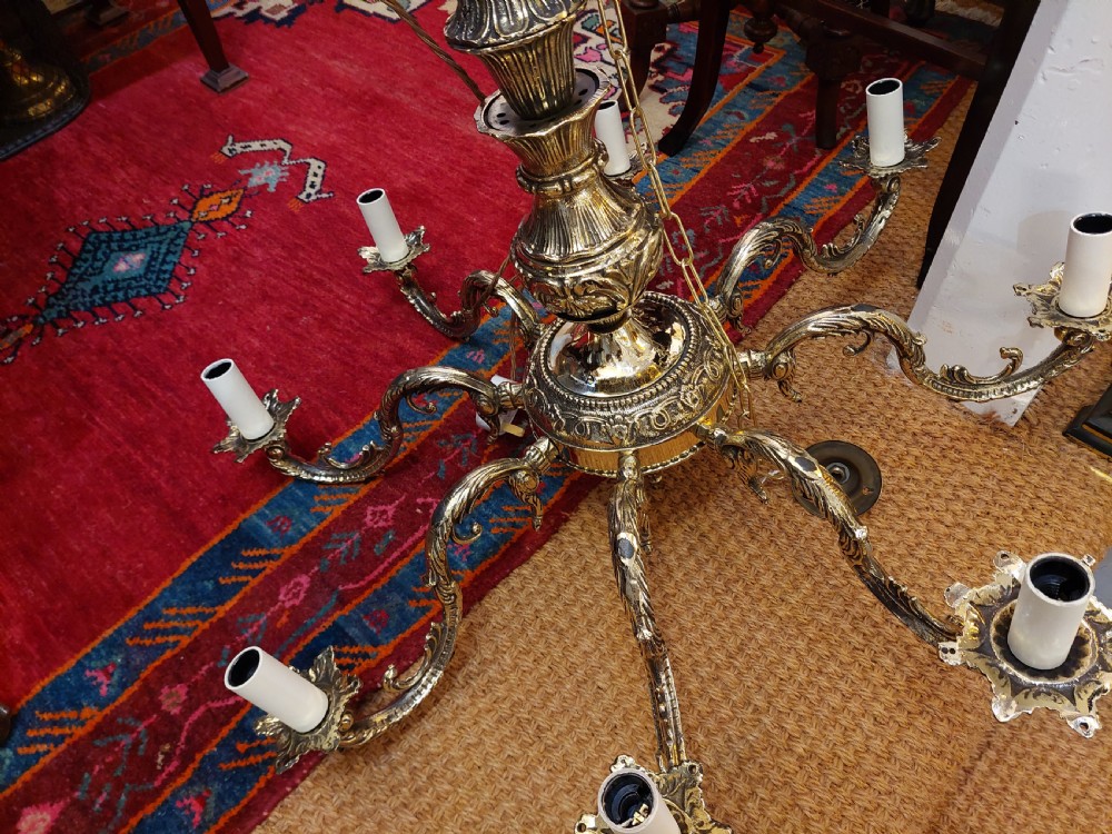 An eight-arm cast brass chandelier featuring detailed acanthus leaf mounts, scrollwork arms, and white candle-effect lamp holders. View X