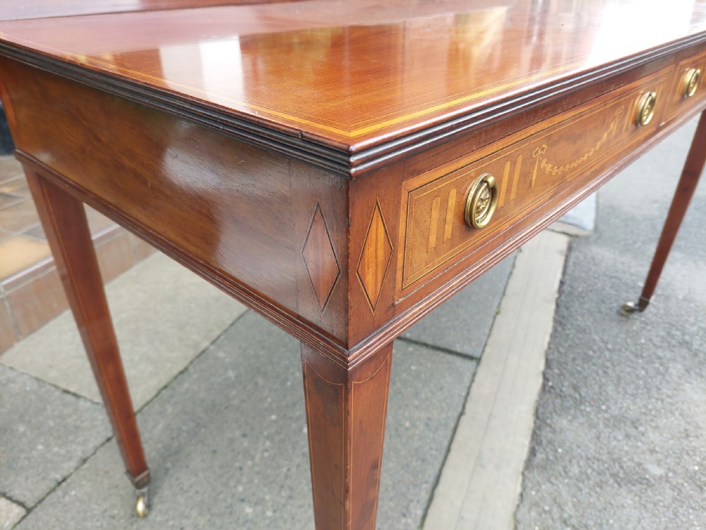 An Edwardian mahogany hall table featuring two inlaid drawers with brass ring handles, tapered square legs with brass castors, and a low rear gallery. View III