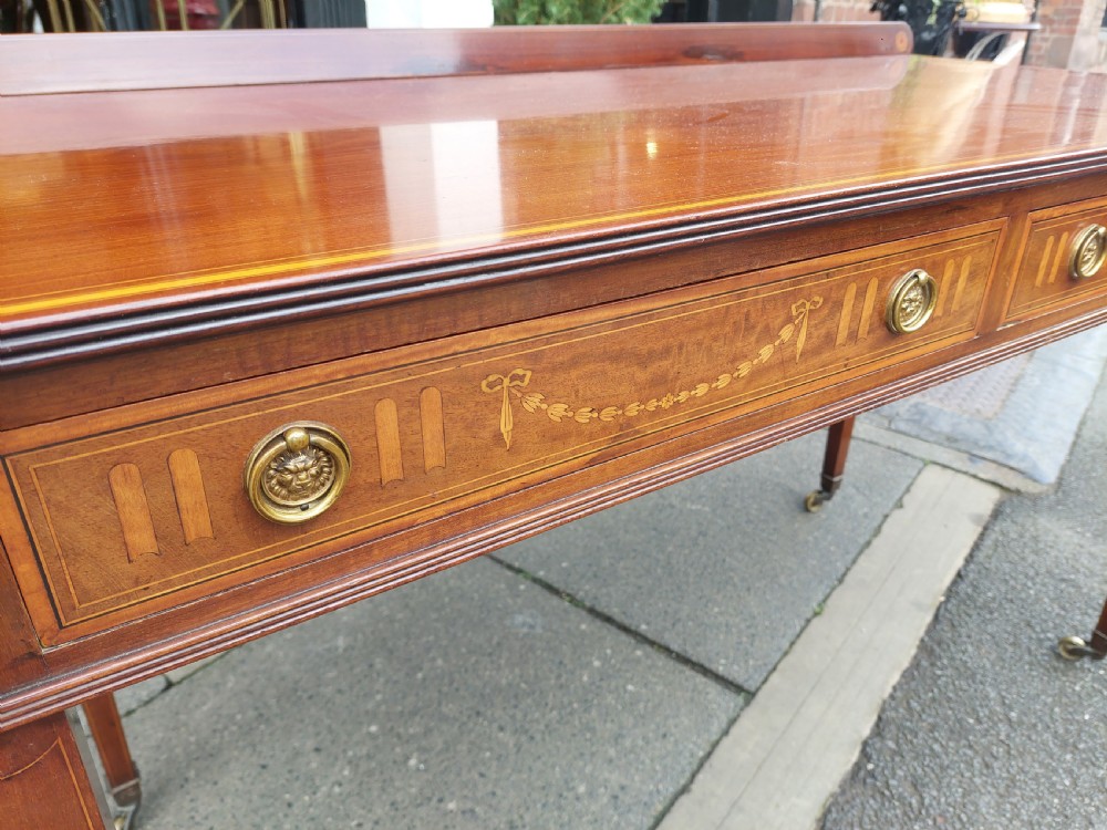 An Edwardian mahogany hall table featuring two inlaid drawers with brass ring handles, tapered square legs with brass castors, and a low rear gallery. View IV
