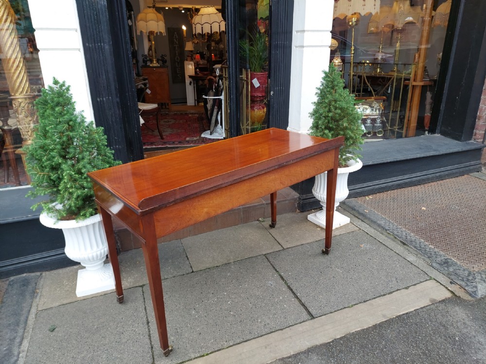 An Edwardian mahogany hall table featuring two inlaid drawers with brass ring handles, tapered square legs with brass castors, and a low rear gallery. View X