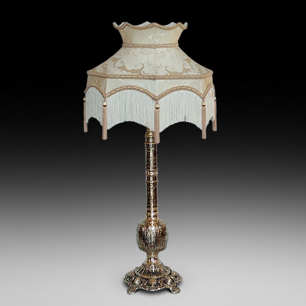 An early 20th-century pressed brass table lamp featuring an ornate relief stem and base, topped with a cream patterned fabric shade with fringe and tassels.