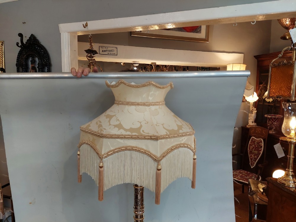 An early 20th-century pressed brass table lamp featuring an ornate relief stem and base, topped with a cream patterned fabric shade with fringe and tassels. View III