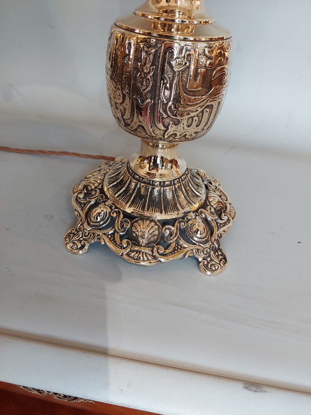 An early 20th-century pressed brass table lamp featuring an ornate relief stem and base, topped with a cream patterned fabric shade with fringe and tassels. View IV