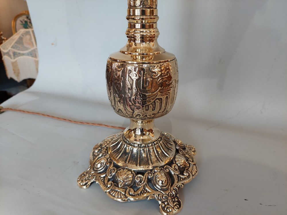 An early 20th-century pressed brass table lamp featuring an ornate relief stem and base, topped with a cream patterned fabric shade with fringe and tassels. View V