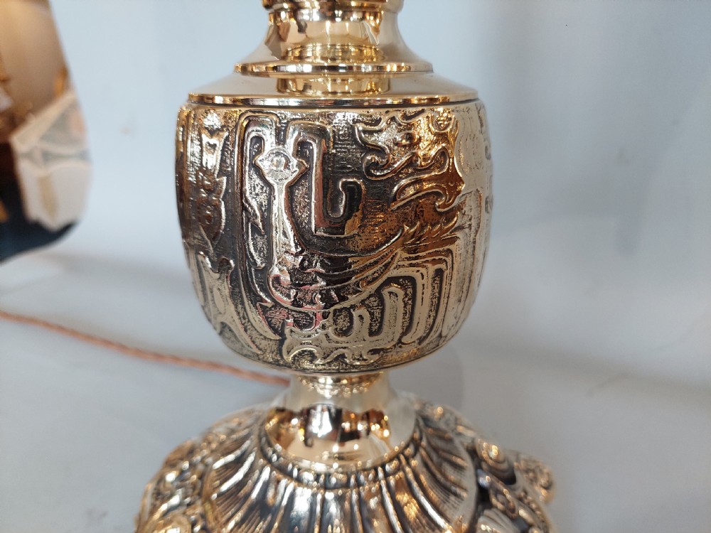 An early 20th-century pressed brass table lamp featuring an ornate relief stem and base, topped with a cream patterned fabric shade with fringe and tassels. View VI
