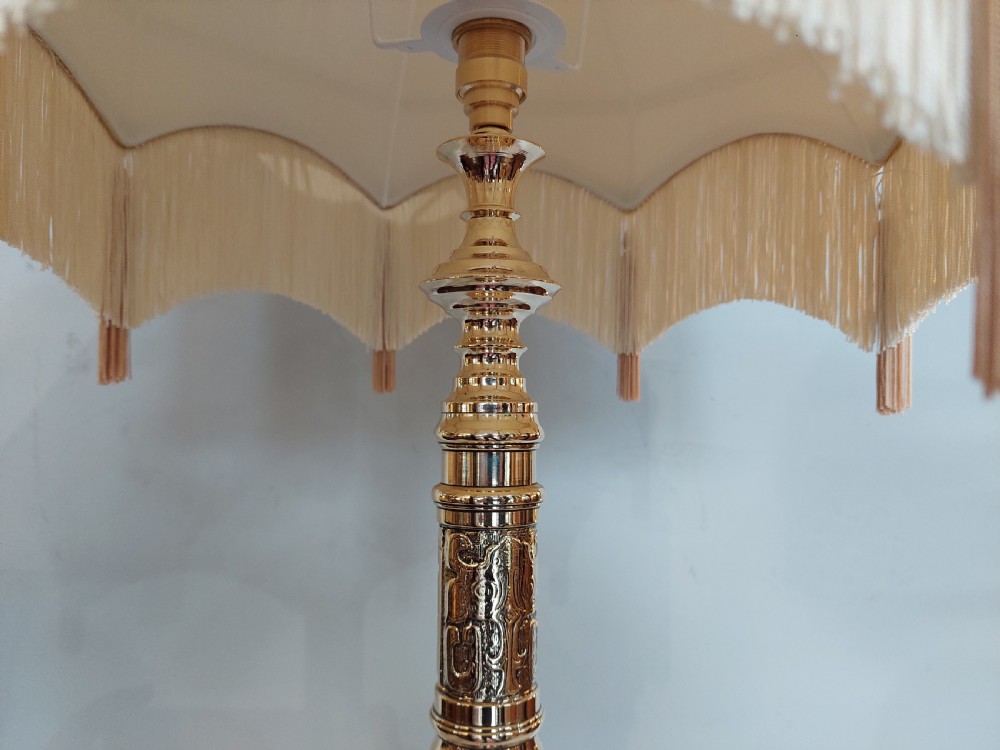 An early 20th-century pressed brass table lamp featuring an ornate relief stem and base, topped with a cream patterned fabric shade with fringe and tassels. View VII