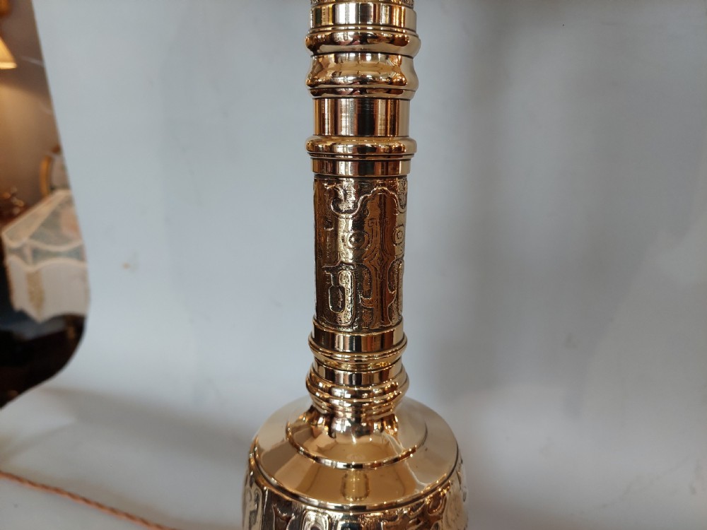 An early 20th-century pressed brass table lamp featuring an ornate relief stem and base, topped with a cream patterned fabric shade with fringe and tassels. View VIII