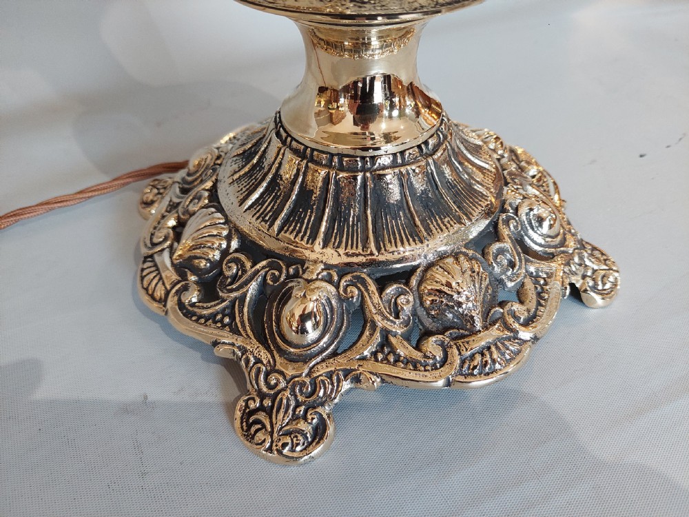 An early 20th-century pressed brass table lamp featuring an ornate relief stem and base, topped with a cream patterned fabric shade with fringe and tassels. View IX