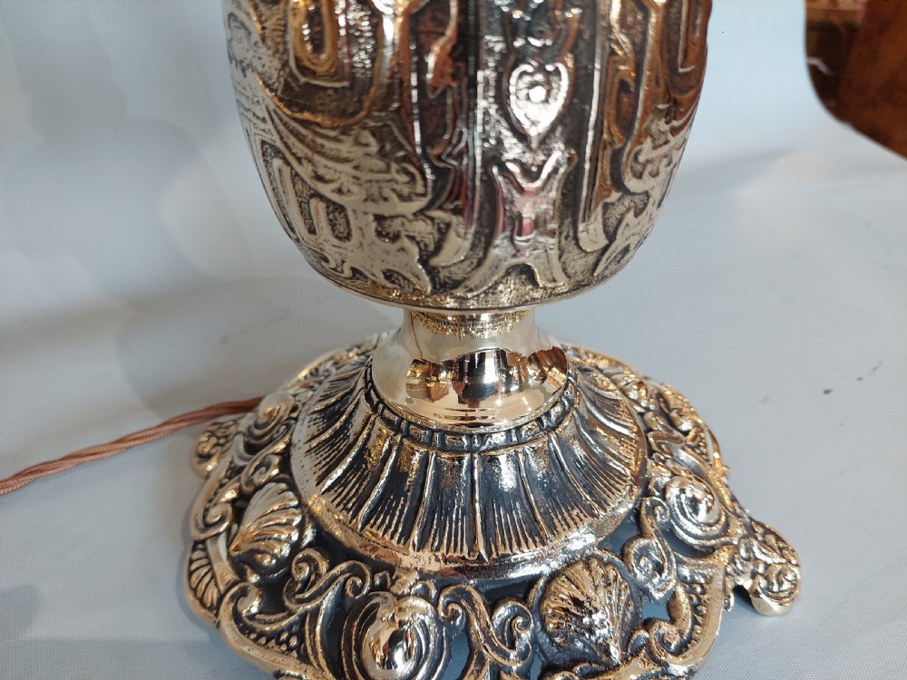 An early 20th-century pressed brass table lamp featuring an ornate relief stem and base, topped with a cream patterned fabric shade with fringe and tassels. View X