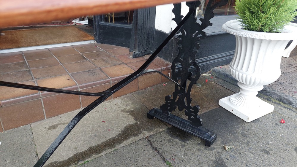A Victorian style rectangular table featuring a polished timber tabletop supported by black ornate cast iron trestle ends and diagonal bracing. View VII