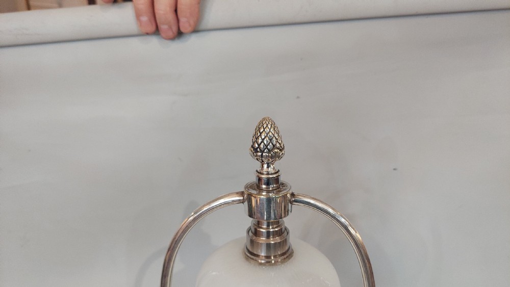 An Edwardian silver-plated table lamp featuring a reeded column, circular gadrooned base, pinecone finial, and an oval hoop frame holding a white frilled glass shade. View IX