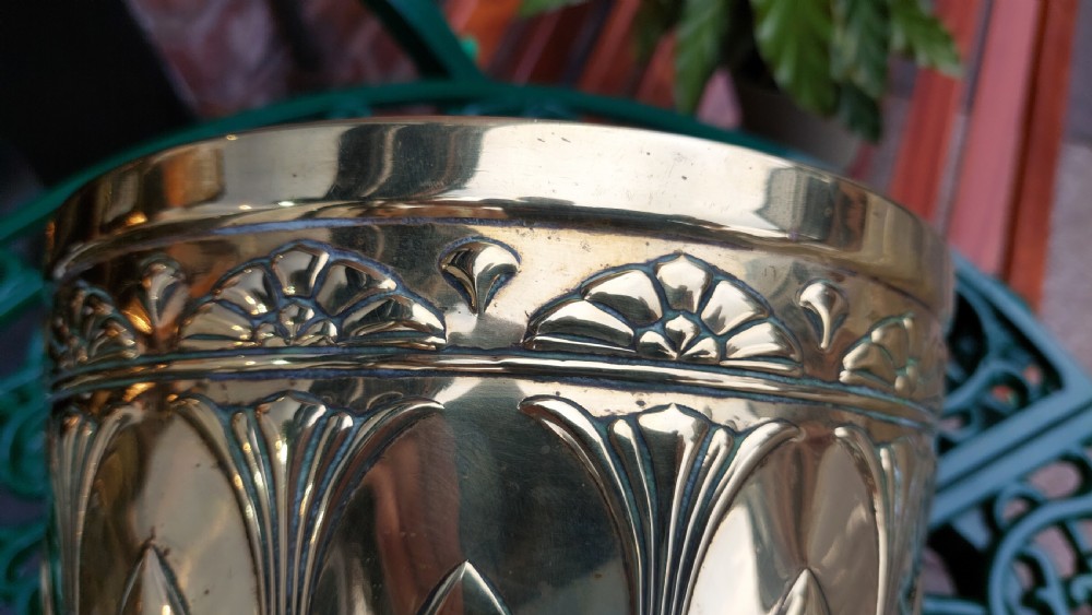 A tapered cylindrical brass planter featuring embossed Art Nouveau floral motifs above a textured hammered base. View V