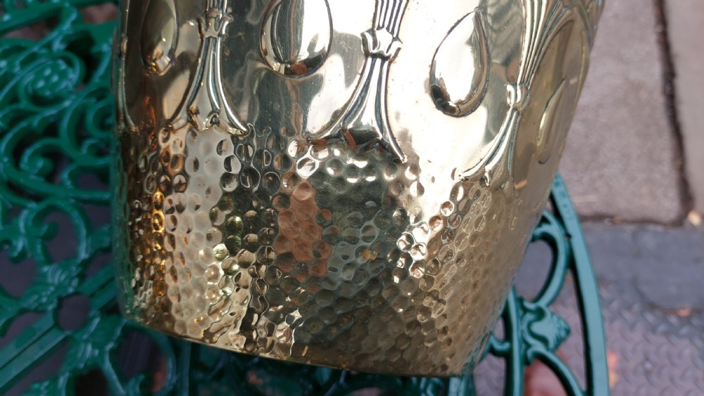A tapered cylindrical brass planter featuring embossed Art Nouveau floral motifs above a textured hammered base. View VII