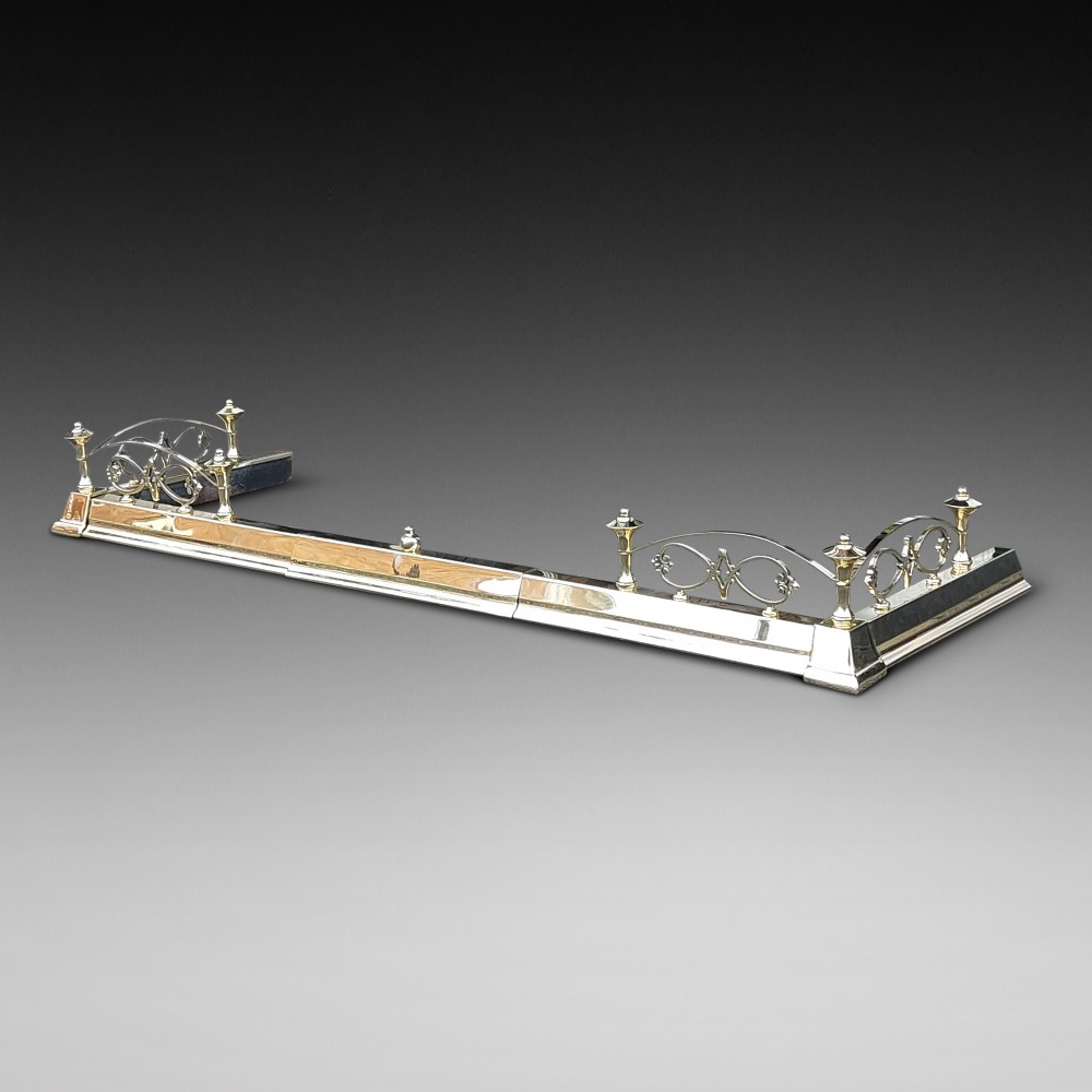 A polished brass extending fireplace fender featuring decorative side gallery rails with scrollwork and urn-shaped corner finials.