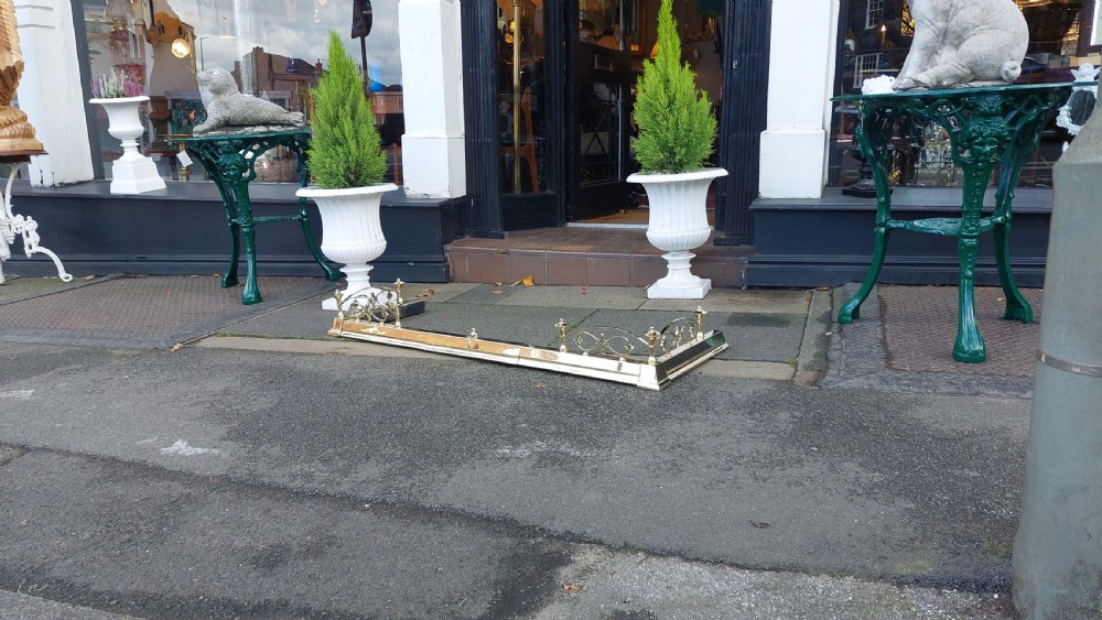 A polished brass extending fireplace fender featuring decorative side gallery rails with scrollwork and urn-shaped corner finials. View III